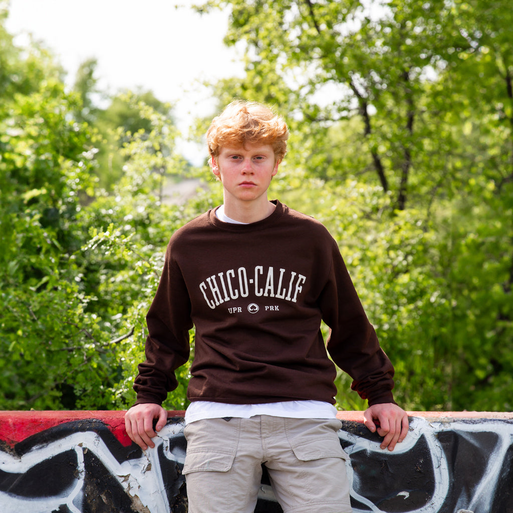 a man sitting on a concrete graffiti barrier wearing a brown Chico Collegiate Thrifty Crew Sweater from Upper Park Clothing in Chico Ca