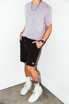 mean leaning against a white wall wearing a orchid simple mineral washed shirt and black men's stadium shorts from Upper Park Clothing