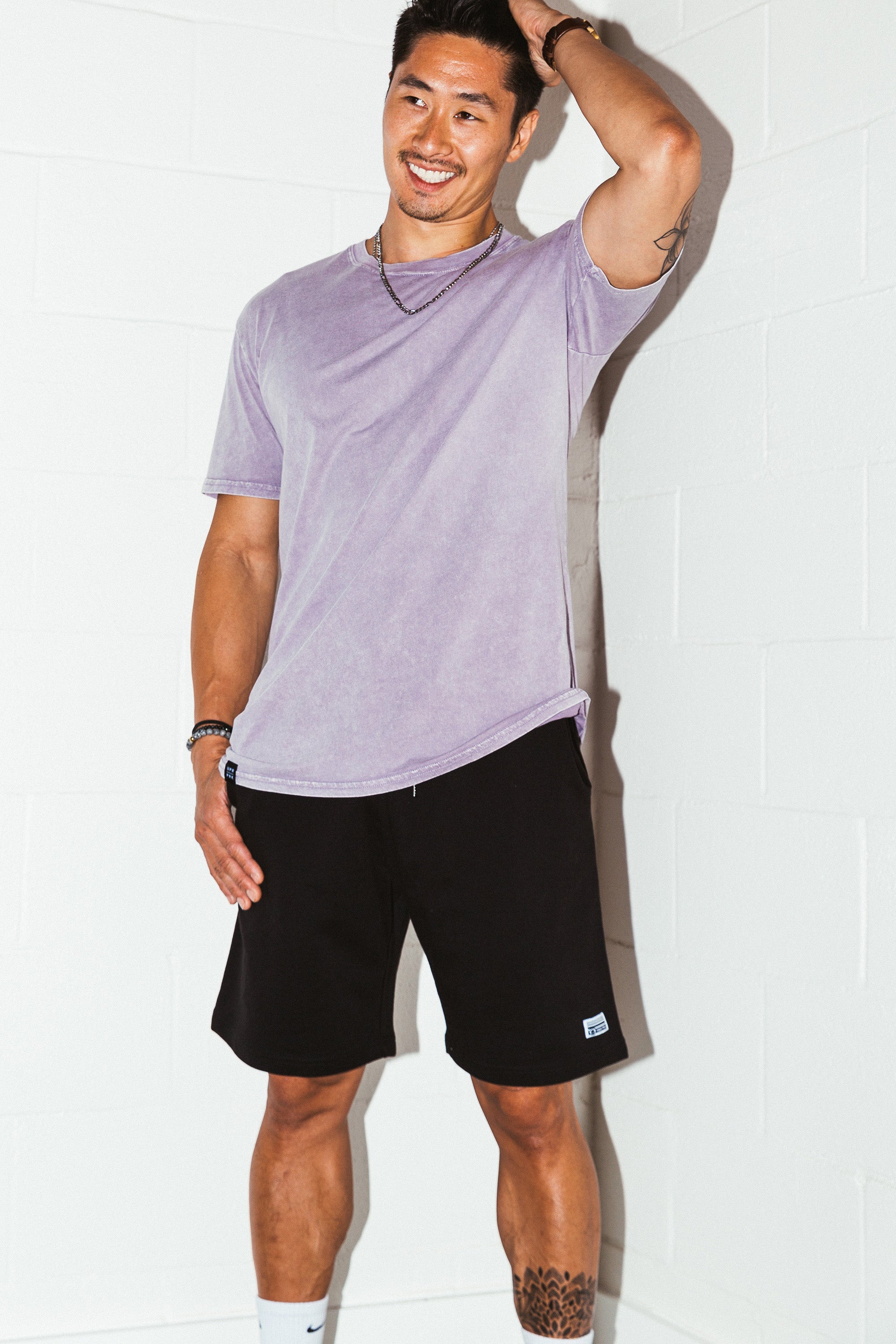 mean leaning against a white wall wearing a orchid simple mineral washed shirt and black men's stadium shorts from Upper Park Clothing