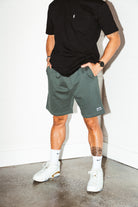 mean leaning against a white wall wearing a black pocket flag tee and green men's stadium shorts from Upper Park Clothing
