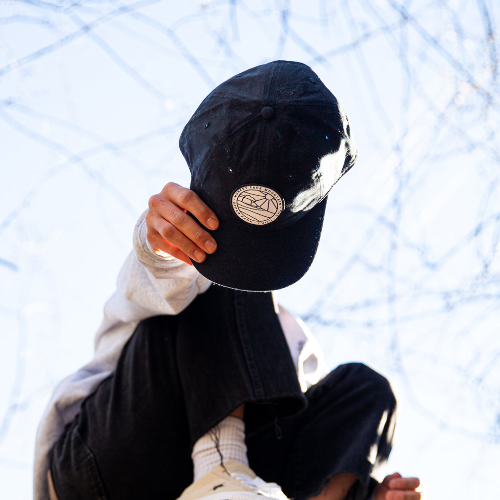 man holding a Monkey Face Mountain Patch Six Panel Black hat in front of his face while sitting in a tree while wearing a California Forests Athletic Heather Crew Neck Sweater from Upper Park clothing in Chico, California