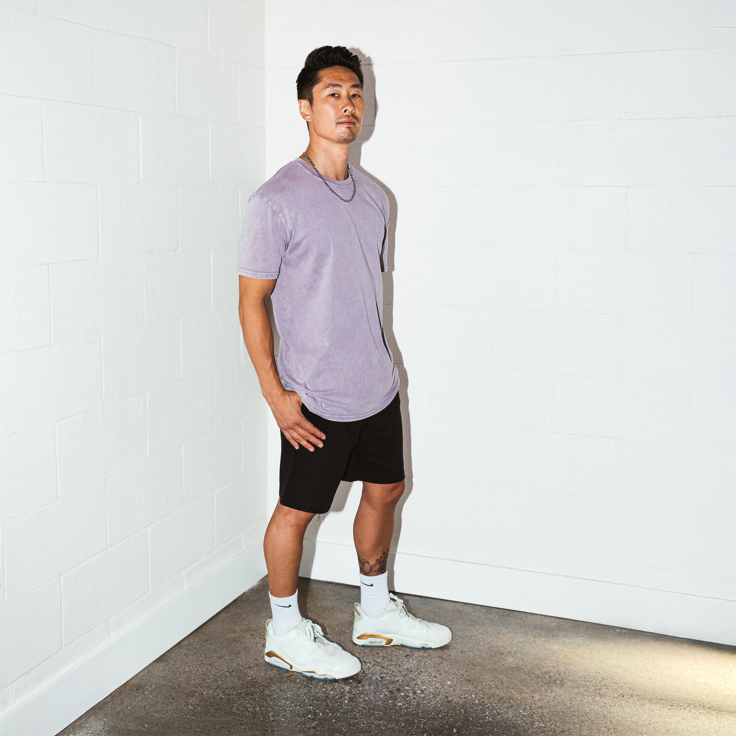 Man wearing a light purple shirt, black shorts, and white sneakers against a white wall. Upper Park Simple Stone Wash Tee