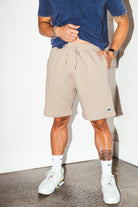 mean leaning against a white wall wearing a blue simple washed mineral tee and tan colored stadium shorts from Upper Park Clothing