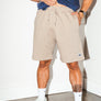 mean leaning against a white wall wearing a blue simple washed mineral tee and tan colored stadium shorts from Upper Park Clothing