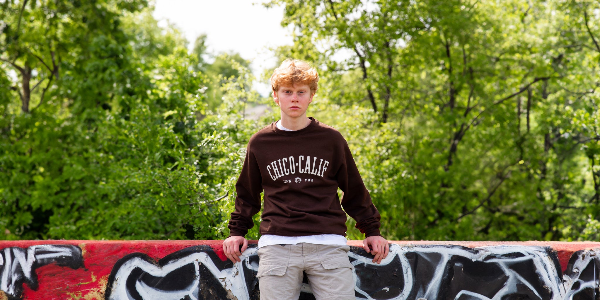 a man sitting on a concrete graffiti barrier wearing a brown Chico Collegiate Thrifty Crew Sweater from Upper Park Clothing in Chico Ca