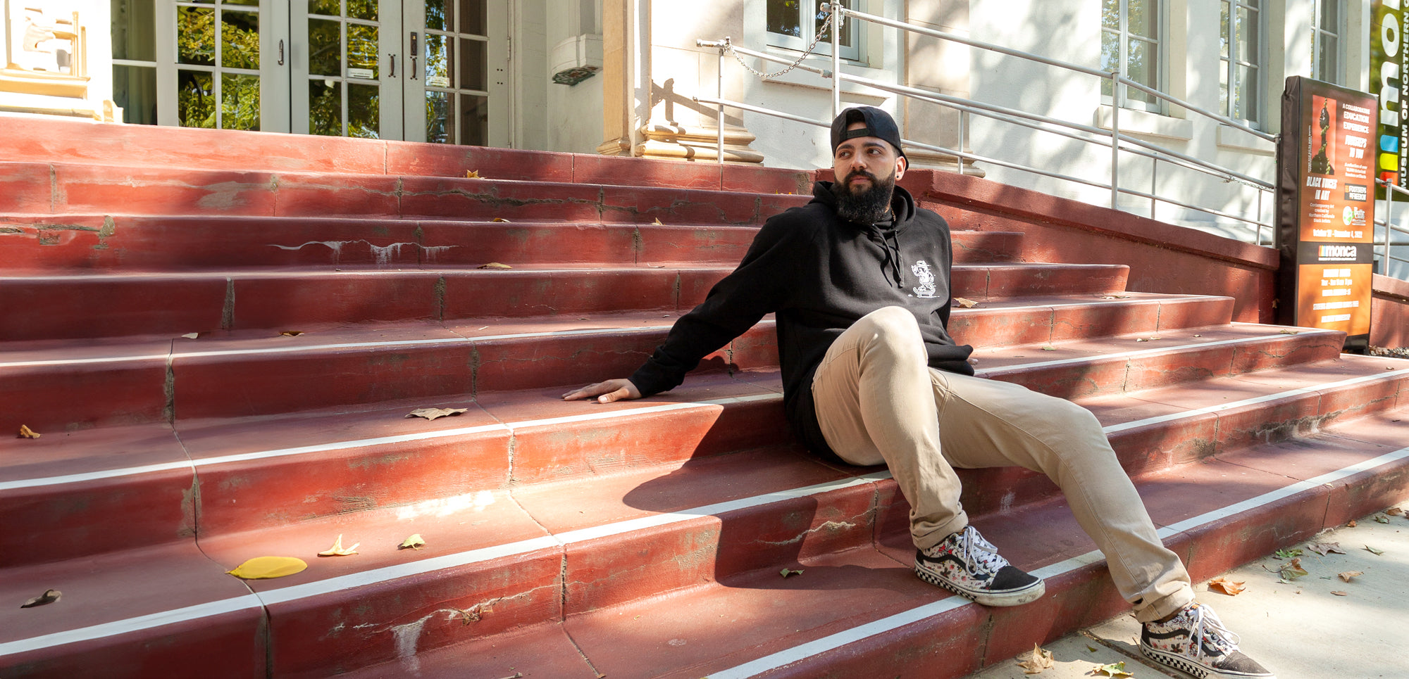 man sitting on a set of stairs in front of MONCA building in Downtown Chico wearing a Corner Store Connoisseurs Hoodie from Upper Park Clothing