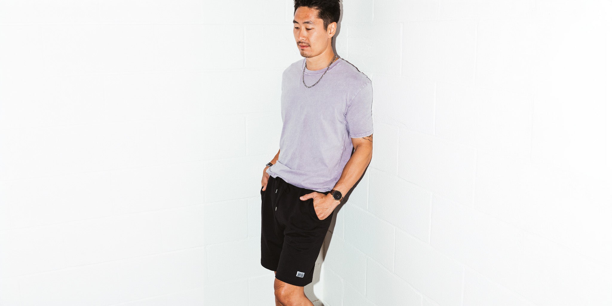 Man leaning against a wall flash photography wearing a Orchid colored simple washed tee and black micro horizon sweat shorts from Upper Park Clothing in Chico California