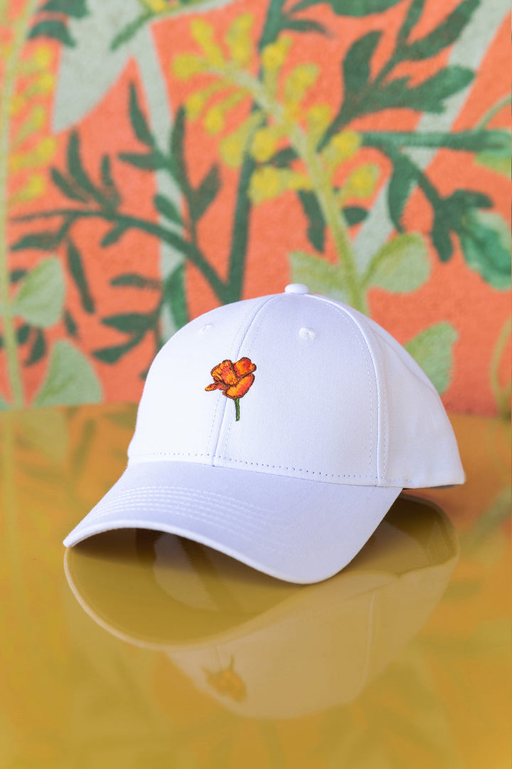 a white California Poppy Hat from Upper Park Clothing sitting on a table in front of a Orange wall with some flowers painted on it