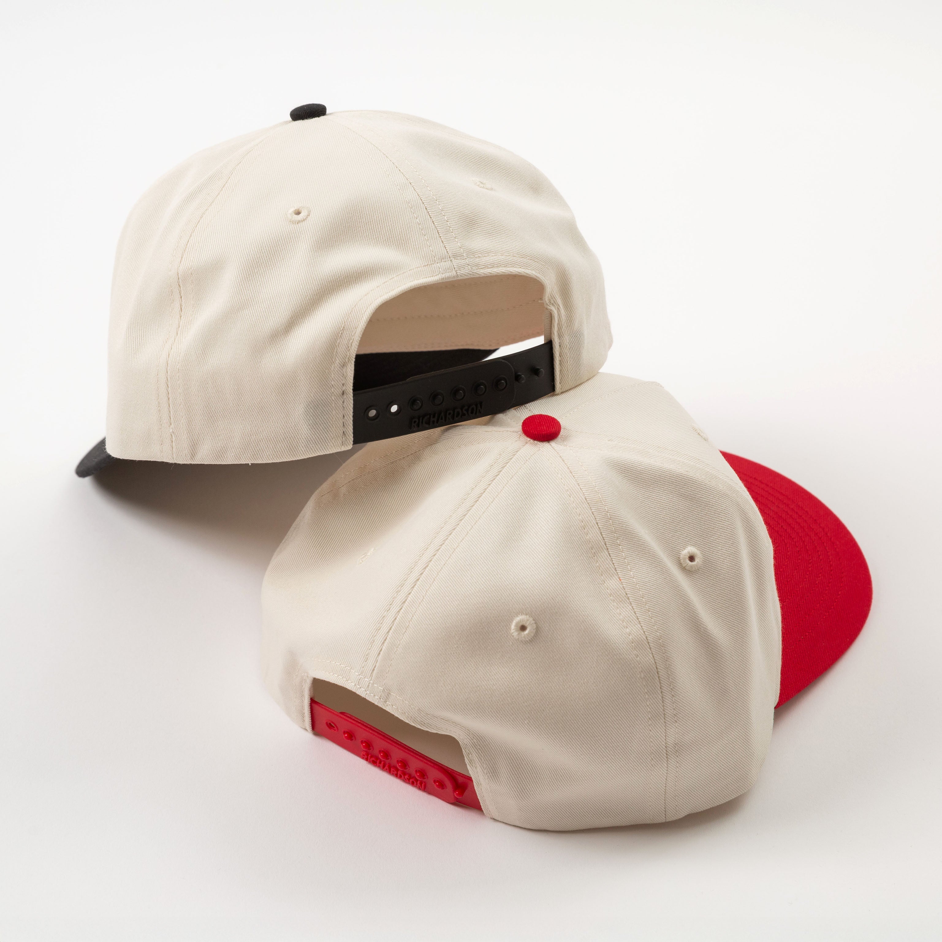 Two vintage style baseball caps, one beige with a black brim and red button, the other beige with a red brim and black button, on a white background.