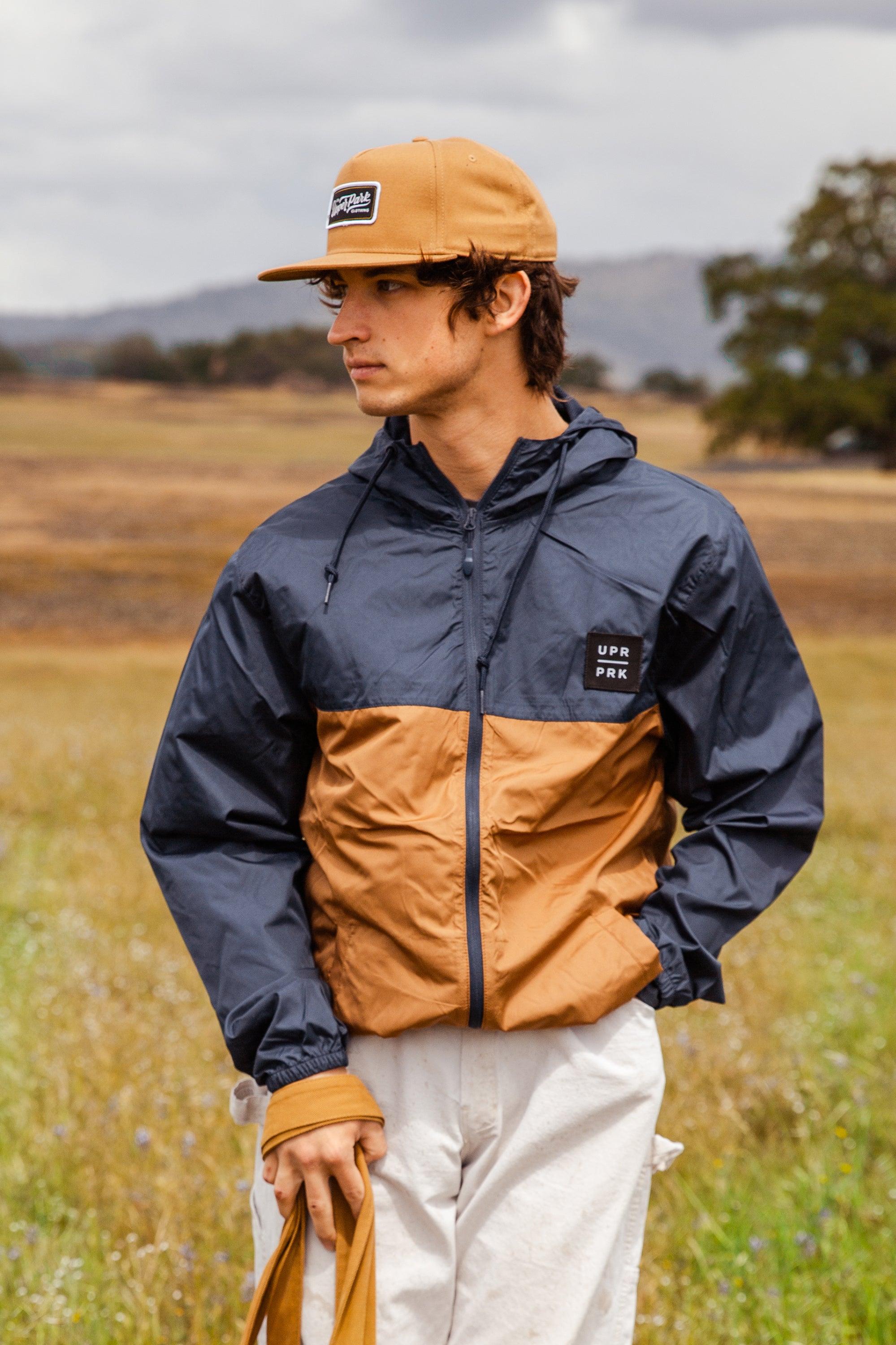 a man wearing a Navy and Saddle colored windbreaker with a Black UPR PRK Patch on the left chest, wearing a Camel colored Workmans Patch Billy Cap Mid Profile hat from Upper Park Clothing in Chico, CA
