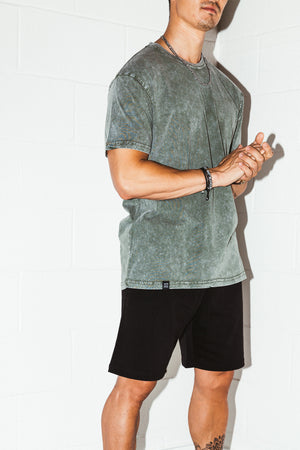 mean leaning against a white wall wearing a green simple mineral washed shirt and black men's stadium shorts from Upper Park Clothing
