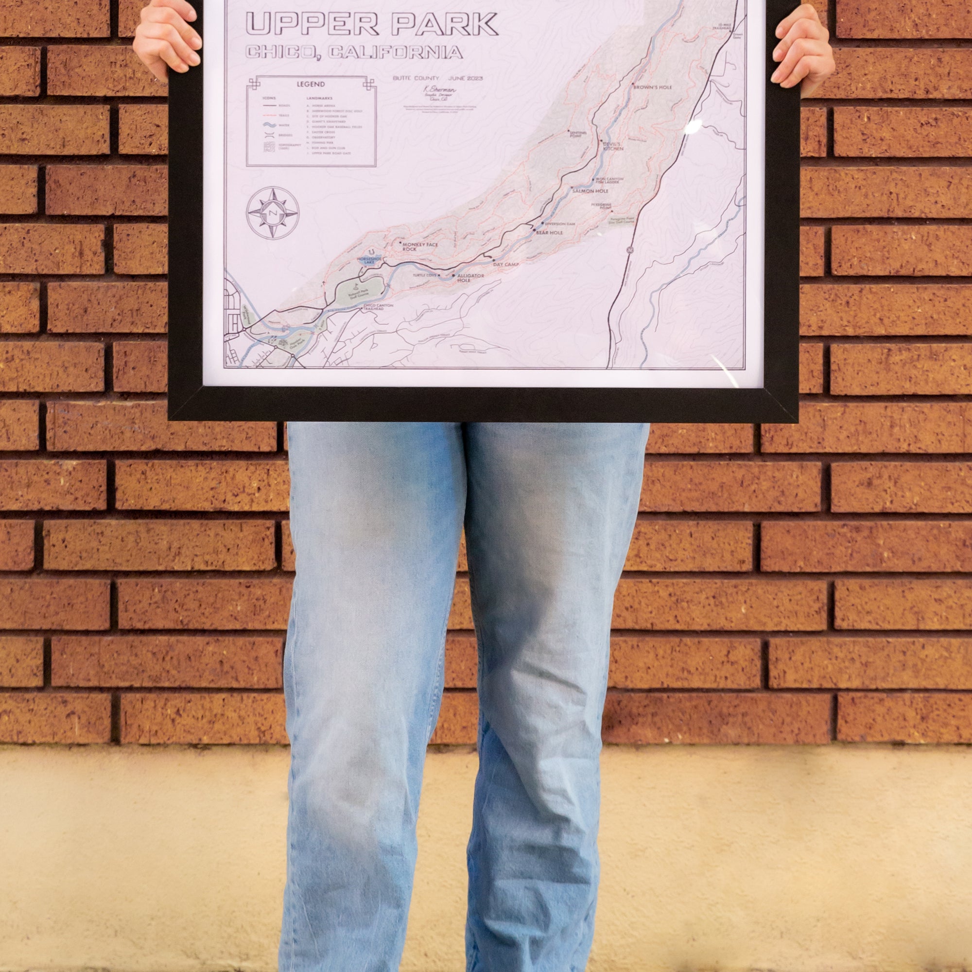 women standing in front of a brick wall in Downtown Chico holding a Upper Bidwell Park Map Poster in a Frame from Upper Park Clothing