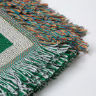 Close-up of a green and white woven fabric with colorful fringe on a light gray background - Upper Bidwell Park Woven Blanket